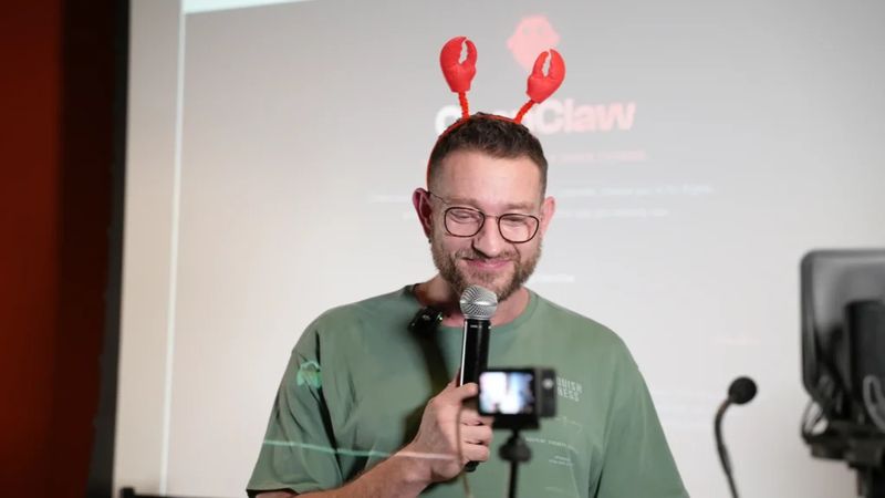 Man wearing lobster claw headband and glasses speaking into a microphone at ClawCon, with the ClawCon logo visible on a screen behind him